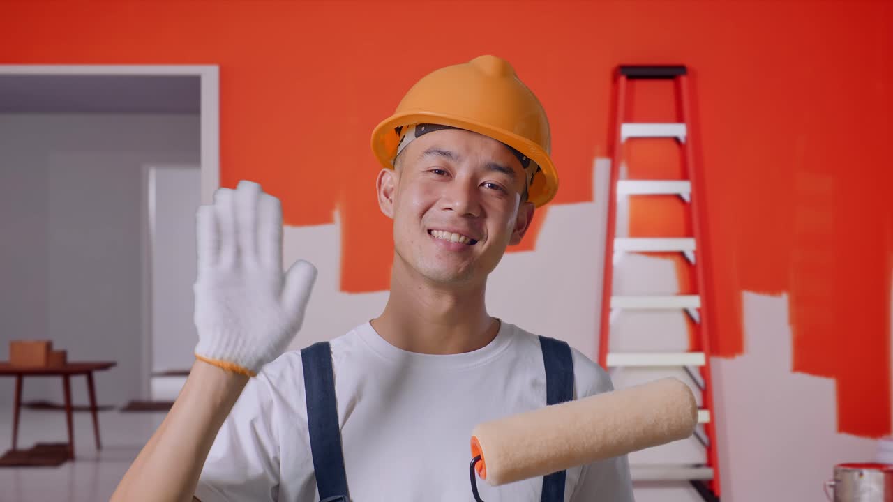 Close Up Of Asian Man Painter Wearing Safety Helmet Smiling, Waving Hand, And Saying Bye While Standing With Painting Wall In Room