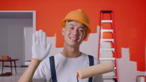 close up de un pintor asiático que usa un casco de seguridad sonriendo, agitando la mano y diciendo adiós mientras está de pie con la pared de pintura en la habitación
