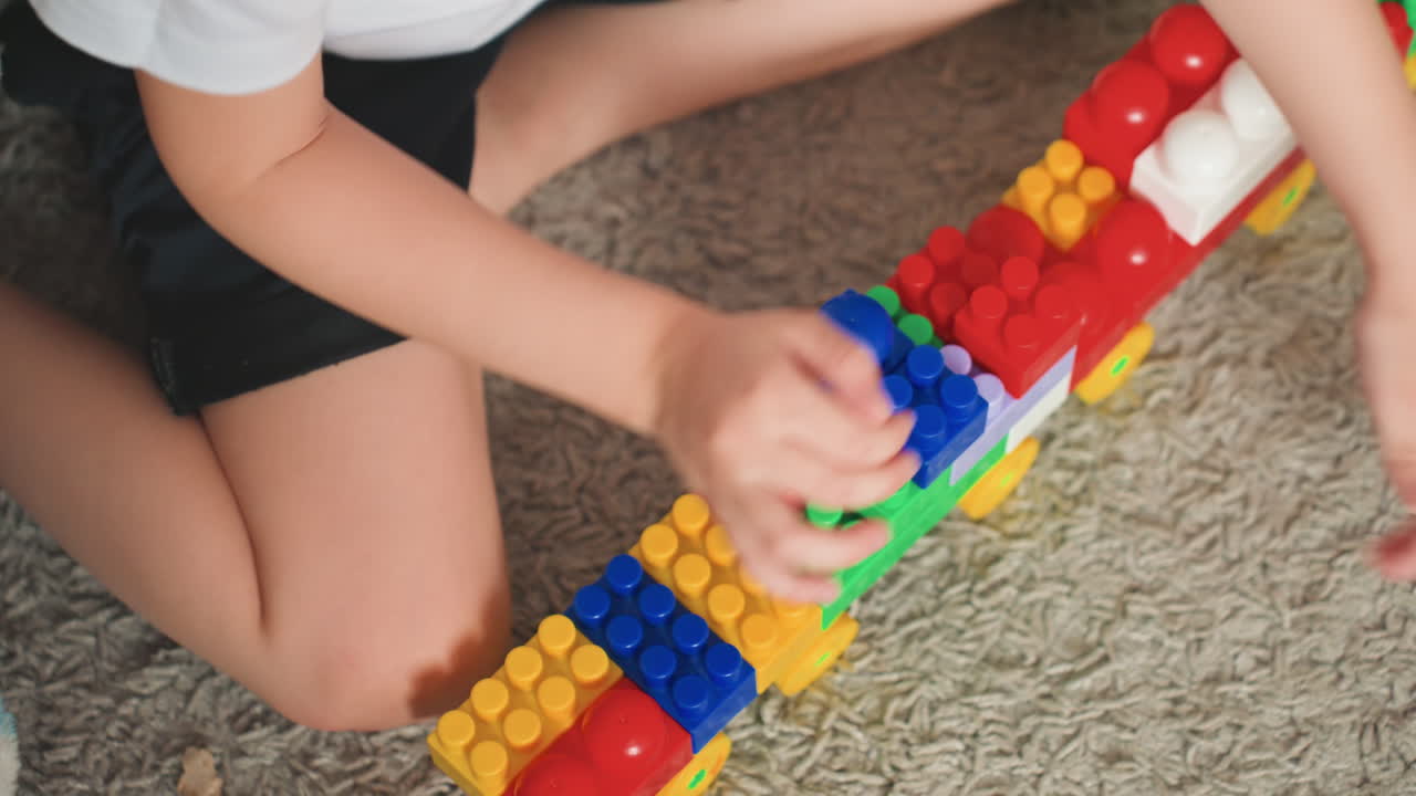Hijo blanco construyendo una larga pared de bloques, joven constructor ensamblando filas coloridas en la alfombra, juego estructural con ladrillos de plástico, creatividad independiente y resolución de problemas, disposición persistente y lúdica.