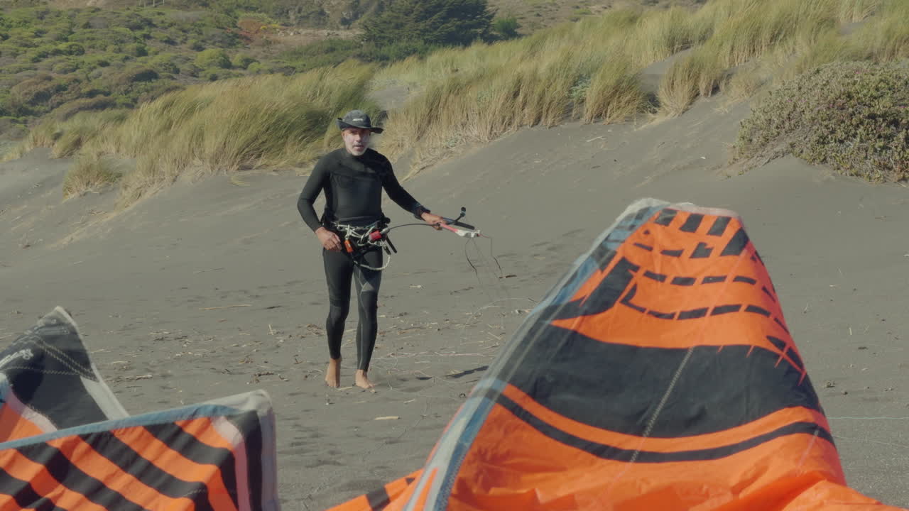 hombre en traje de buceo comprobando la línea de aparejo mientras camina de regreso a orange kitesurf vela en la playa ventosa en matanzas, chile