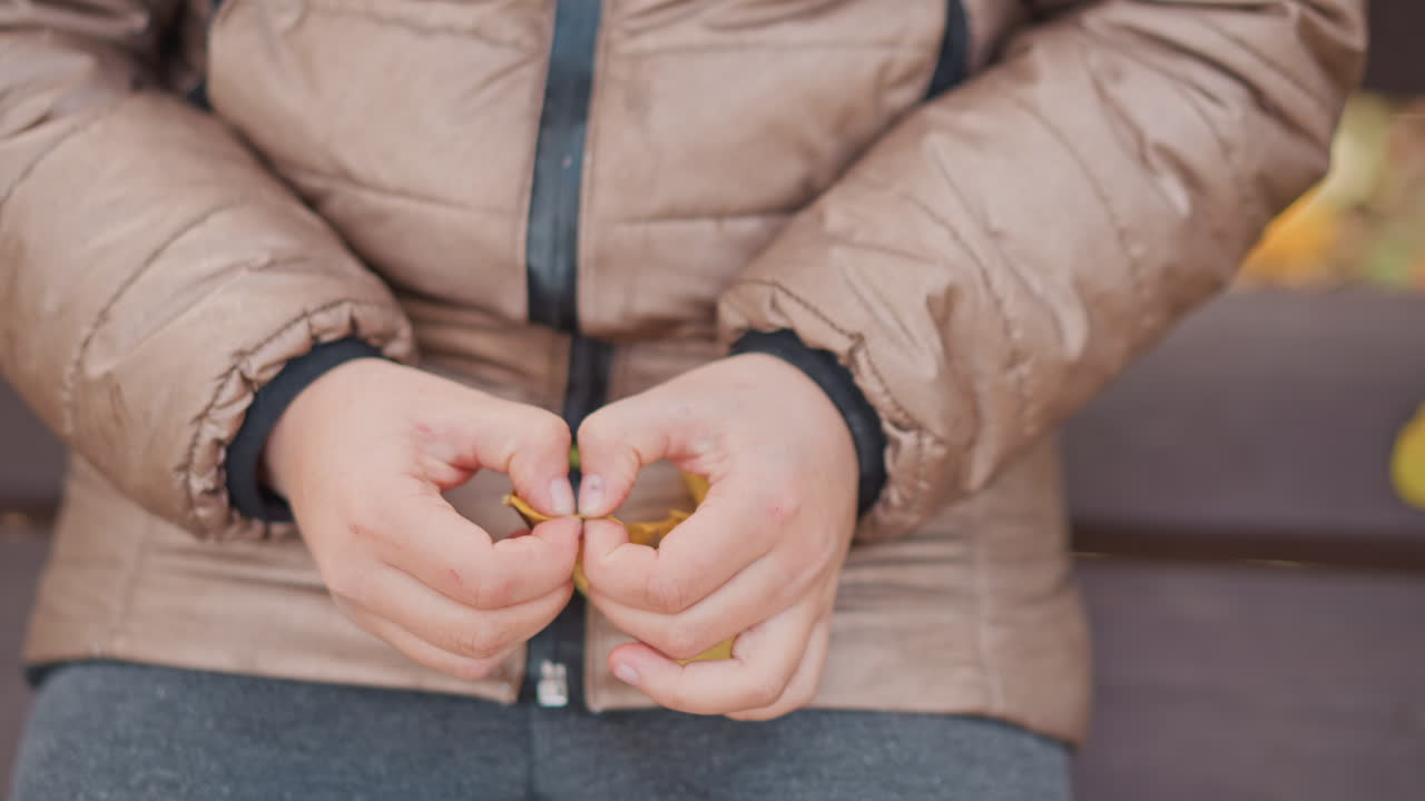 close up of child in puffy jacket and gray sweatpants seated on ornate wooden bench, careful fingers peeling and tracing yellow autumn leaf veins, blurred second leaf nearby