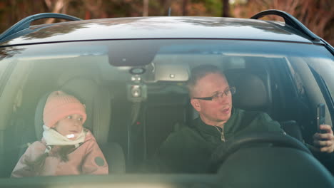 a man is holding a smartphone, with a young girl sitting beside him in the front passenger seat,the man, wearing glasses and a green jacket, the girl, dressed in a pink cap and jacket, looks up at him