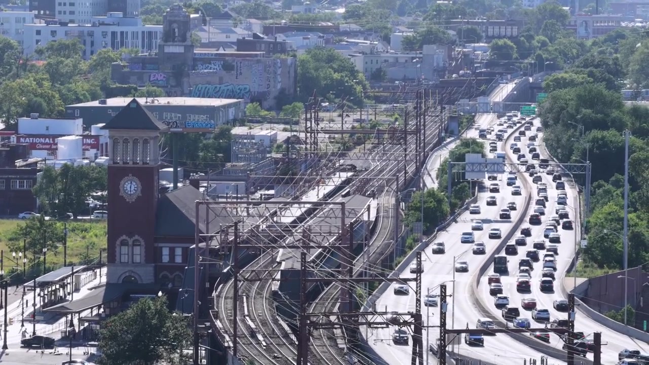Una storica torre dell'orologio in mattoni si erge accanto ai binari della ferrovia e a un treno merci di passaggio a Newark, New Jersey, con un intenso traffico autostradale che si accumula lungo un'autostrada curva a più corsie. Ampia ripresa aerea con drone.