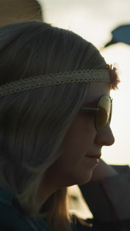 un primer plano de una mujer con una cinta de flores en la cabeza y gafas de sol, ajustando sus gafas mientras está sentada en un campo de hierba, alcanza una hebra de hierba