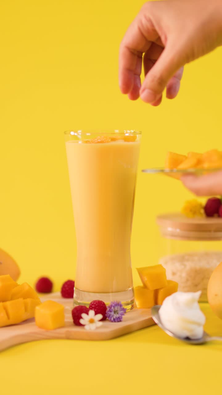 Una mano umana decora un frullato di mango alto con cubetti di frutta su uno sfondo giallo brillante in studio