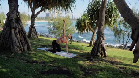 yogi on yoga mat exercising and doing the kneeling twist pose - lifting one arm up and look up towards her outstretched hand - burleigh heads beach in gold coast, qld, australia