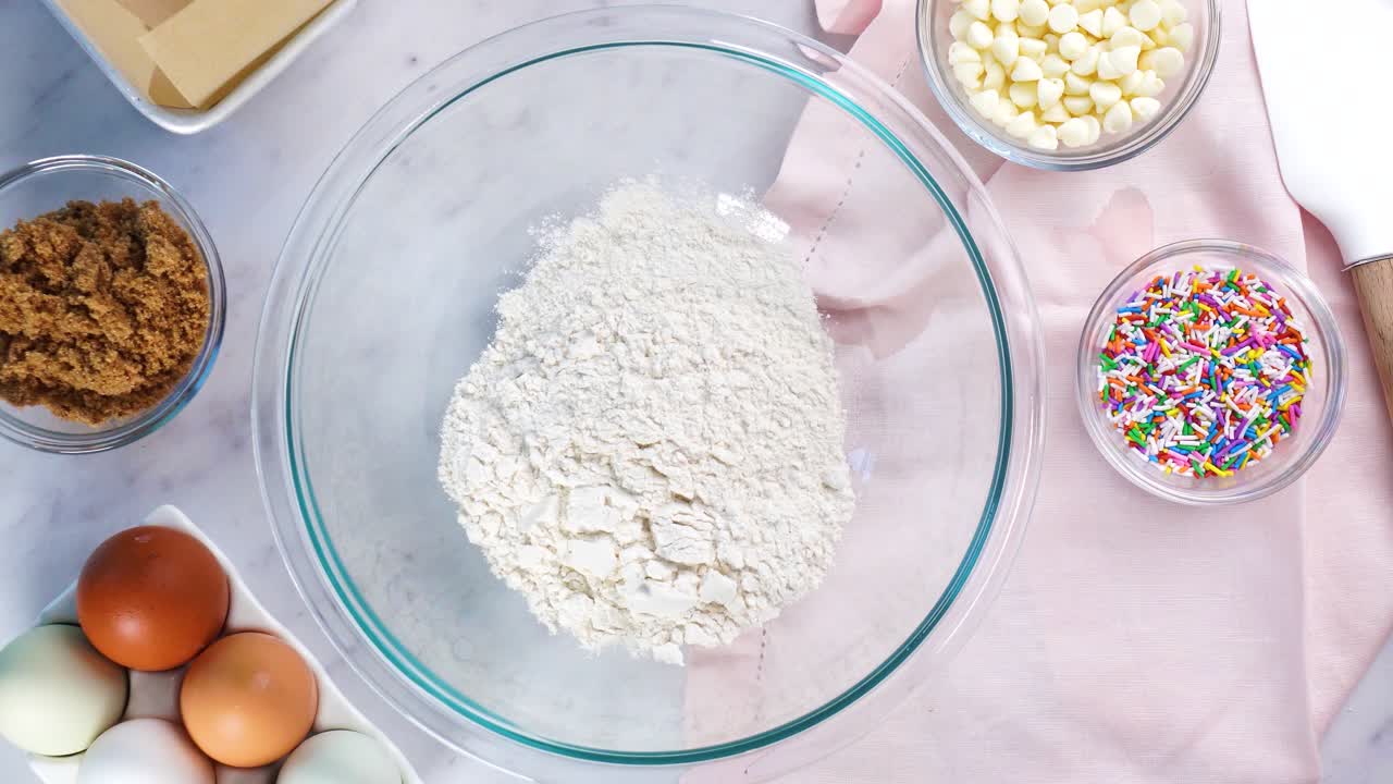 Baking confetti sugar cookies and using a white electric mixer and adding butter to the batter. In the kitchen making a dessert. Making confetti cookies with a white background, pink napkin and eggs