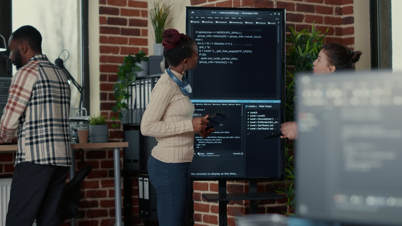 Programmer holding digital tablet analyzing code on wall screen tv while colleague is looking for errors