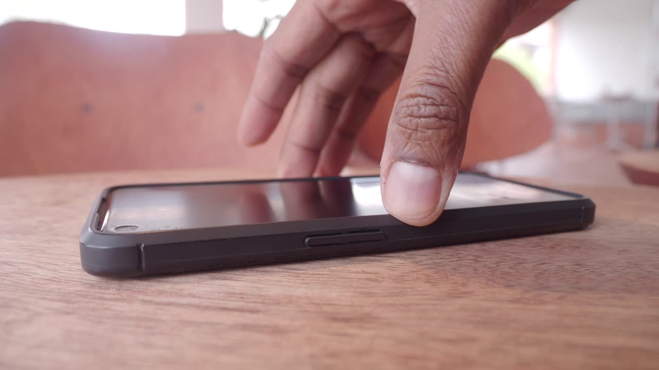 A smartphone on a wooden table with a hand and chairs in the background