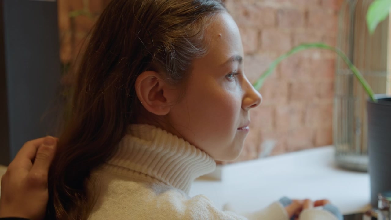 Close up of woman with long brown hair wearing white turtleneck another woman in plaid gently touching her back and neck. Soft lighting highlights natural texture details as they sit