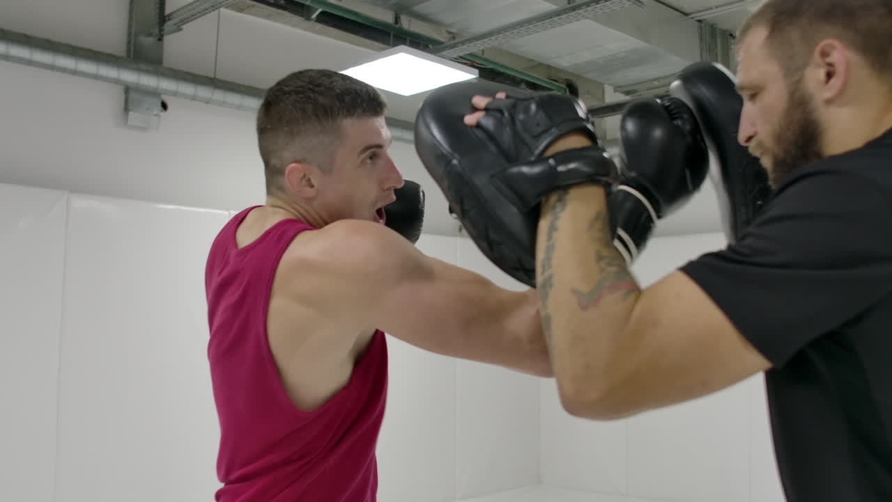 백색 방에서 권투 훈련 세션에서 한 남자가 그의 장갑을 입은 손을 코치의 발바닥에 치고, 타격에서 벗어나고 다이빙을 수행합니다.