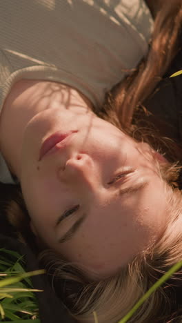 joven acostada pacíficamente en un campo cubierto de hierba, rodeada de altas hojas verdes de herba, con la luz del sol proyectando sombras suaves en su rostro mientras parpadea sus ojos suavemente