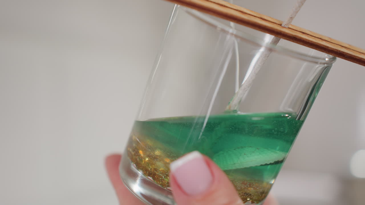 Close-up of glass cup containing wick, golden beads, and seashell with green liquid poured into it, showcasing detailed candle crafting process, liquid settling on bottom