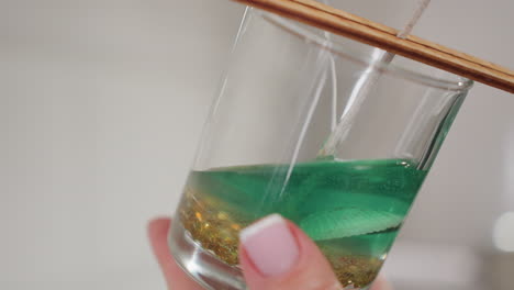 close-up of glass cup containing wick, golden beads, and seashell with green liquid poured into it, showcasing detailed candle crafting process, liquid settling on bottom