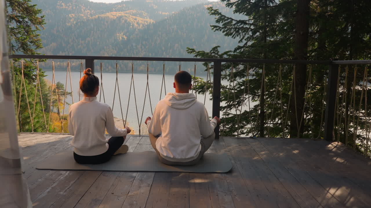 молодая пара медитирует на коврике на террасе. сосредоточенные люди делают упражнения йоги для расслабления вместе, глядя на озеро. концепция осознанного образа жизни