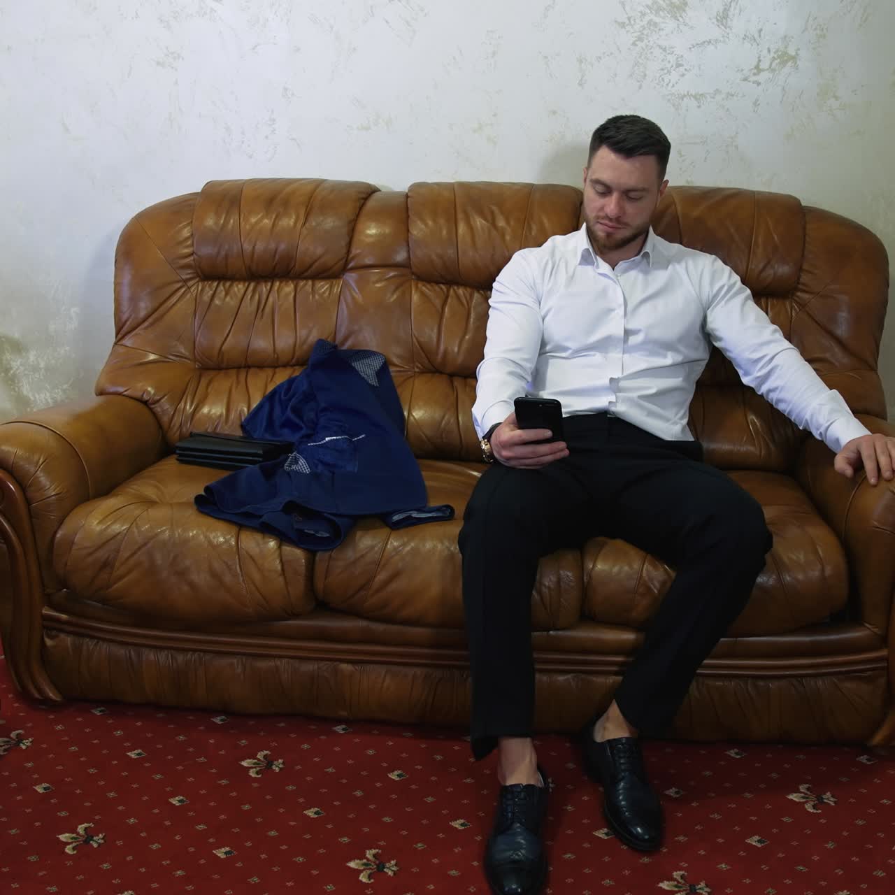 Tired businessman is resting in hotel room. Young man is sitting on sofa and looks at mobile. Comfortable trip away from home