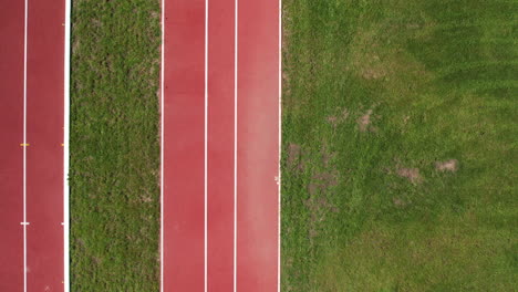 Drohne-Schwenkt-Von-Oben-Nach-Unten-über-Leeres-Rotes-Gleisgummi,-Gesäumt-Von-Gemähtem-Grasfeld