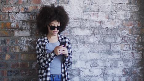 Afroamerikanische-Frau-Mit-Sonnenbrille-Und-Smartphone,-Während-Sie-Gegen-Eine-Ziegelwand-Steht