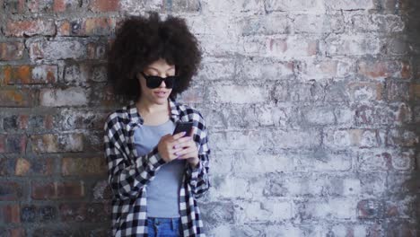 mujer afroamericana que usa gafas de sol usando un teléfono inteligente mientras está de pie contra una pared de ladrillo