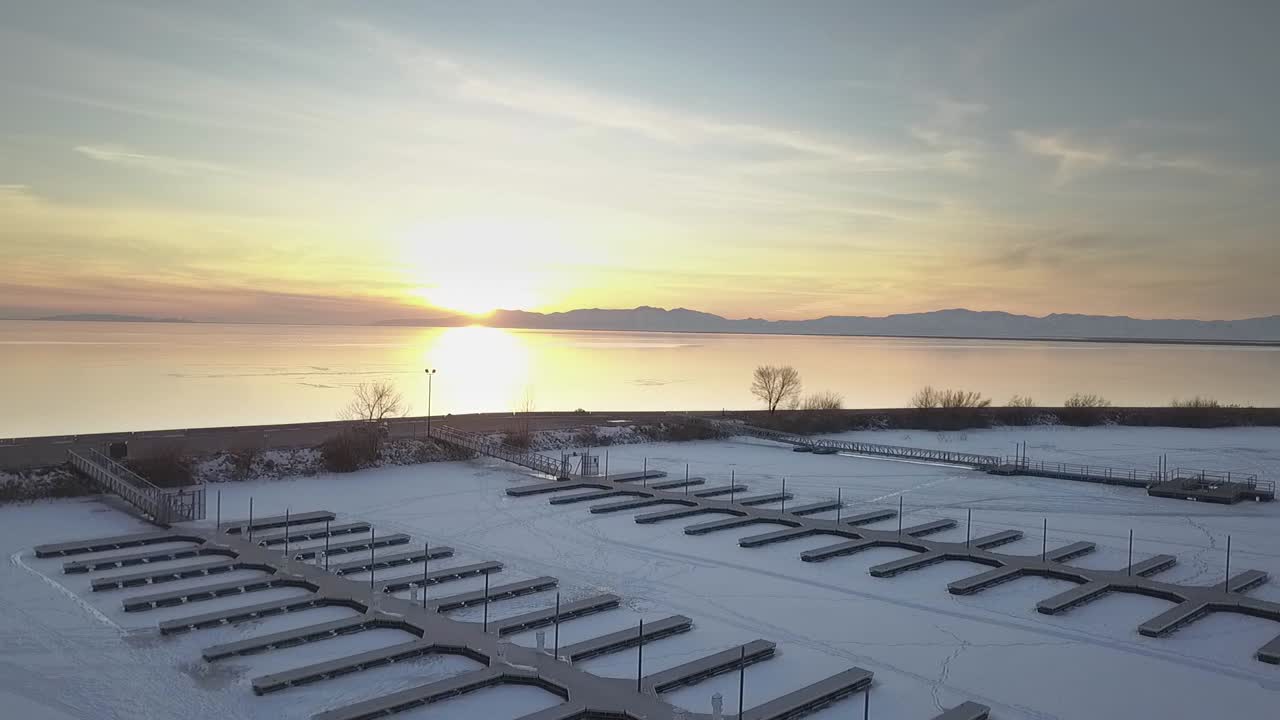 Drone shot flying over frozen lake covered in ice and snow flying with mountains and sunset reflecting off water going downward towards boating dock