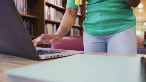 una estudiante asiática con un hijab amarillo sentada en un escritorio sosteniendo un libro y usando una computadora portátil