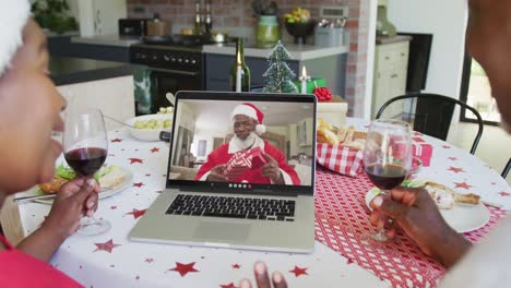 African-american-couple-with-wine-using-laptop-for-christmas-video-call-with-happy-santa-on-screen