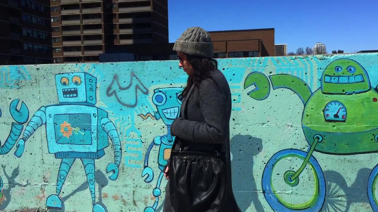 Inquadratura a campo medio di una donna con berretto e cappotto invernale, che si sistema gli occhiali da sole mentre passa accanto a un murale d'arte pubblica in una giornata di sole.