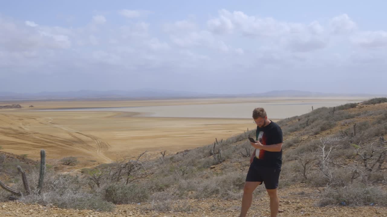 Young caucasian man walks up the desert hill checking his phone and overlooking the scenic landscape of Guajira, Colombia.