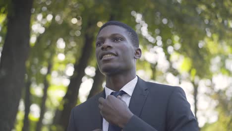 close-up of young handsome african american man adjusting necktie in summer park and leaving. portrait of confident businessman walking for meeting. business, lifestyle, confidence, success.