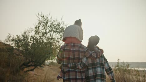 Rear-view-of-a-happy-family,-a-brunette-girl-in-a-hat-in-a-green-checkered-shirt-walks-with-her-husband,-a-man-in-a-brown-checkered-shirt,-and-their-blonde-daughter-in-a-white-jacket-sits-on-the-man’s-shoulders-during-their-hike-and-walk-outside-the-city