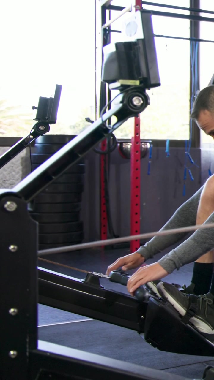 Vertical video: Entering man in shorts and top adjusting straps and rowing erg at gym training