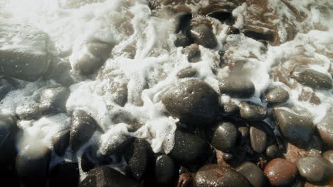 black round small wet pebbles on a stony beach violently washed by ocean wave on a sunny day close up in slow motion