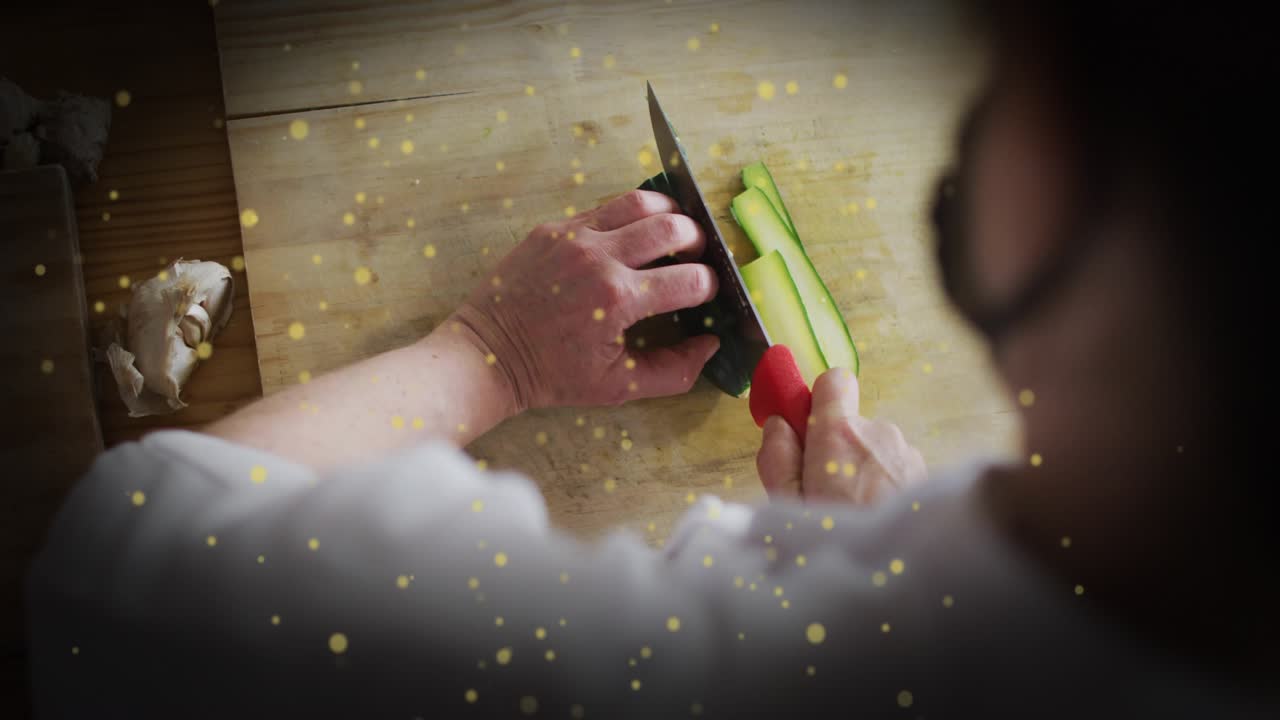 Chef gripping red handled knife slicing cucumber strips for culinary prep, with golden particles