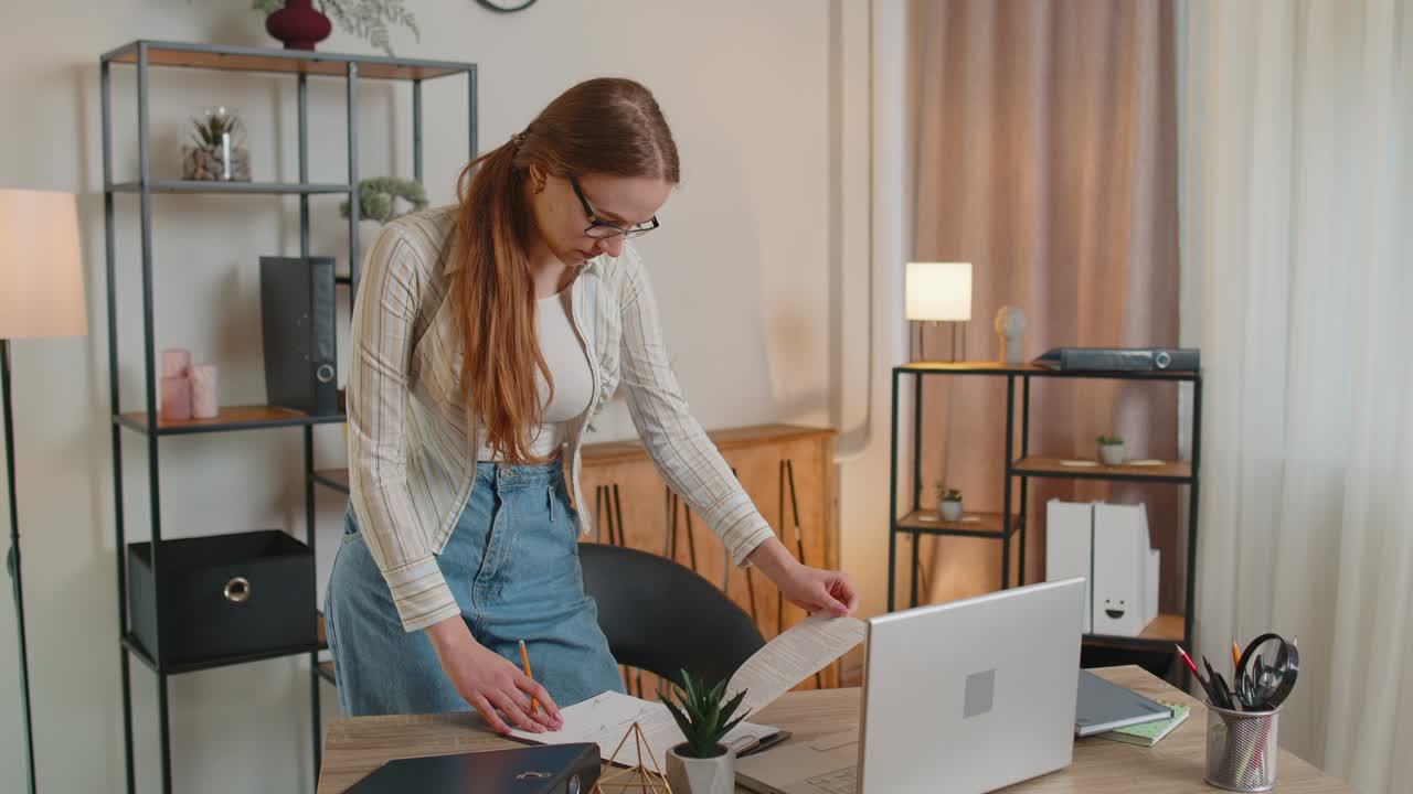 Young woman girl standing by desk in home office, working at paper documents analyzes checks data