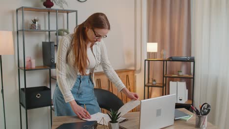 young woman girl standing by desk in home office, working at paper documents analyzes checks data