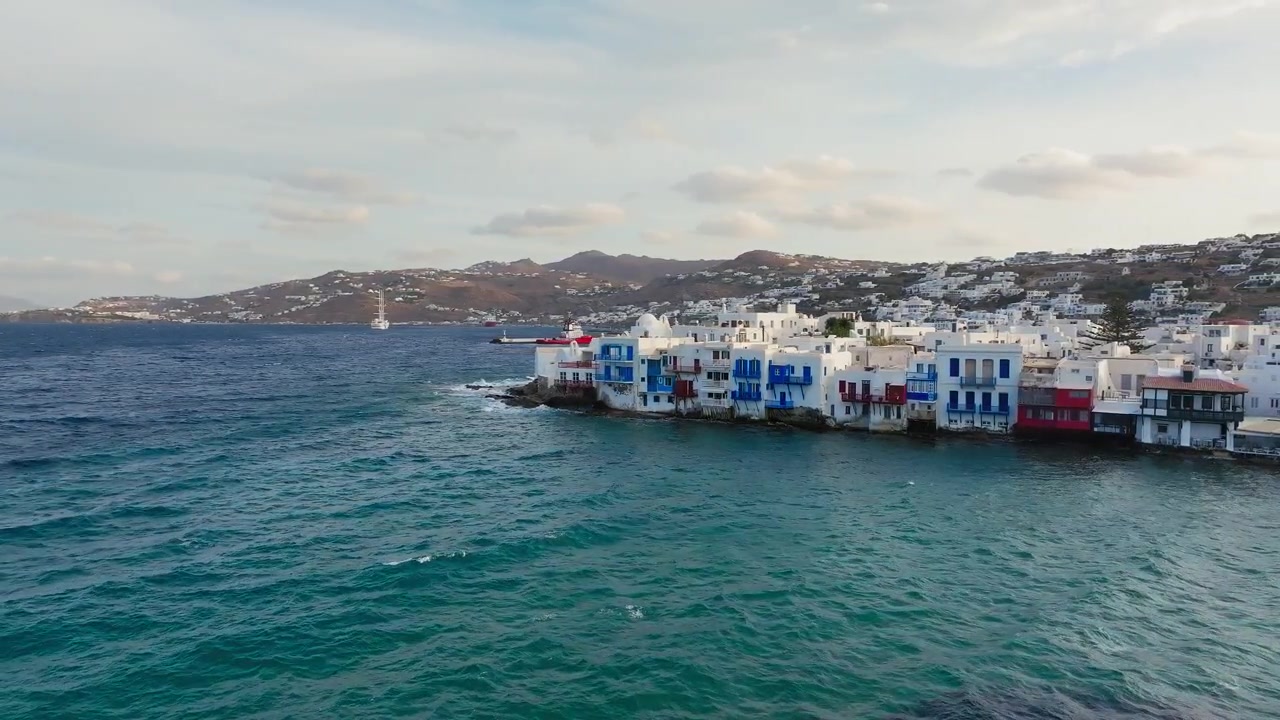 Lenta ripresa panoramica con drone di Piccola Venezia, isola di Mykonos all'alba, Grecia. Edifici colorati sul lungomare, nuvole sparse, luce dorata del mattino.