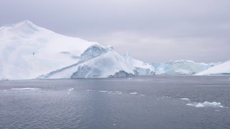 slow motion pan of a large iceberg with small floes of ice in front of it - floating on a calm ocean under a cloudy sky off the coast of greenland