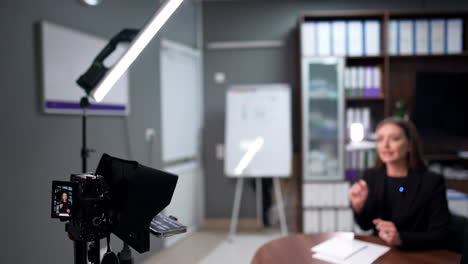 Female educator shooting video for her students. Brunette Caucasian lady in black jacket gives a lesson in front of camera. Woman speaks distinctly and gestures with her hands. Blurred backdrop.