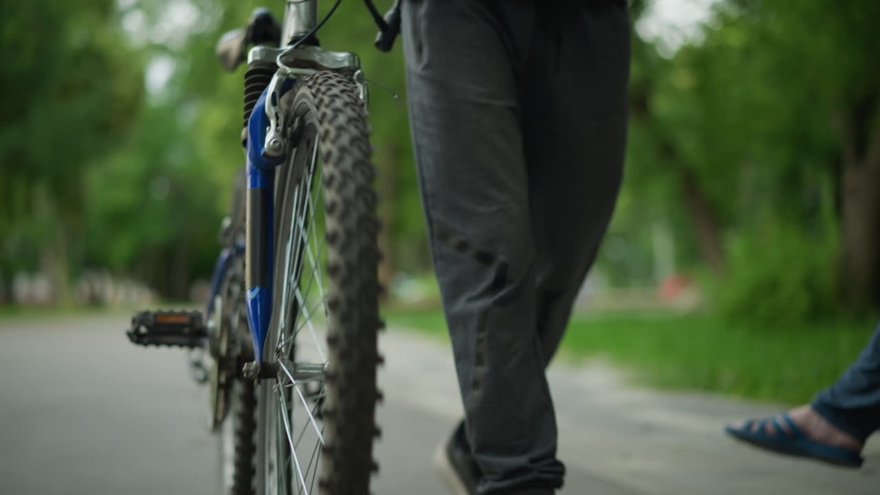 vista parcial de alguien en la parte superior negra y pantalones grises caminando de cerca con bicicleta a lo largo de un camino tranquilo, con vista borrosa de la vegetación y la dama sentada cerca en la distancia