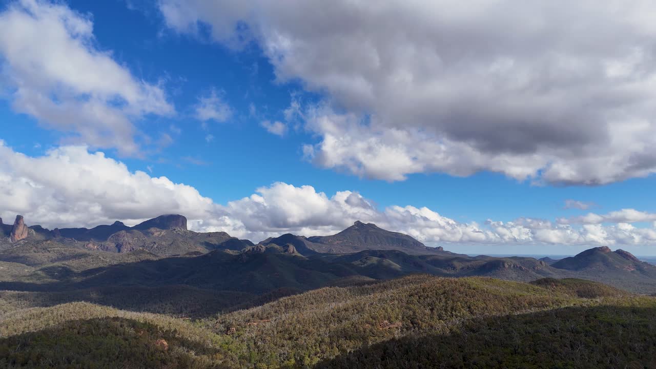 La telecamera di un drone esegue una panoramica fluida attraverso l'aspro paesaggio montuoso di Warrumbungle sotto nuvole drammatiche e la luce del sole del tardo pomeriggio, rivelando la vasta natura selvaggia australiana con dettagli vividi.