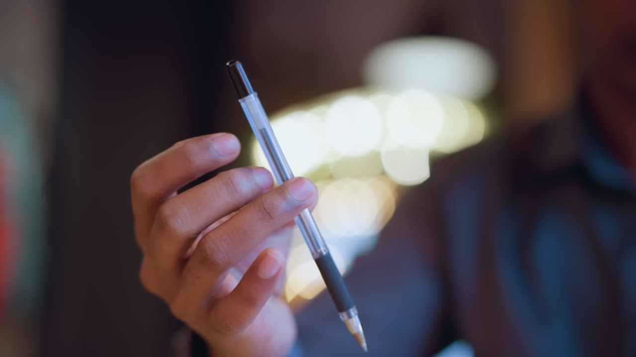 Close-up of male hand gracefully holding pen between fingers in soft indoor lighting with blurred bokeh background, conveying mood of thought, decision-making, communication, or professional expression