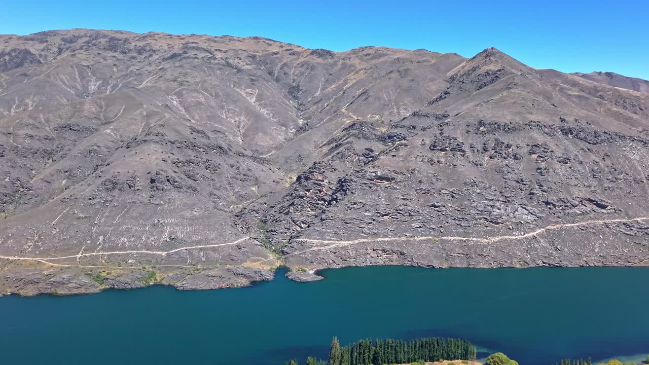 A sunny day drone shot begins with the Dunstan Trail bridge in view, then moves backward to reveal the surrounding mountains and rugged Central Otago landscape