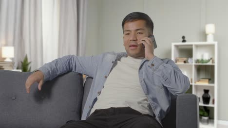 young man sitting on sofa at home smiling and talking with friend on mobile phone