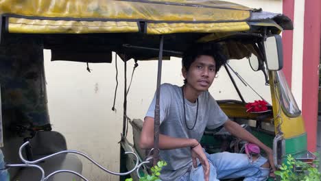 an young indian auto driver in grey t-shirt sitting inside the auto looking at the camera