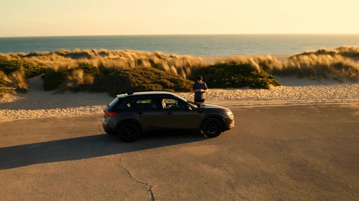 Dark compact SUV sits beside wind-swept coastal dunes at sunset while single man stands near driver's side checking smartphone on quiet paved road with calm ocean horizon and warm golden light