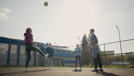 coach leads training session with three ladies playing volleyball on outdoor court, ball drops to ground as coach reaches out to retrieve it with building and fence in background