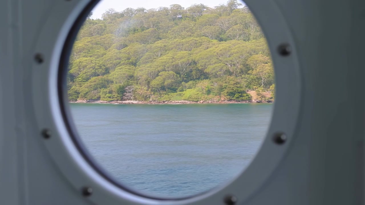Bullauge eines Schiffes rahmt Küstenwald und Meer ein, ruhiges türkisfarbenes Wasser entlang der baumgesäumten Küste sichtbar.