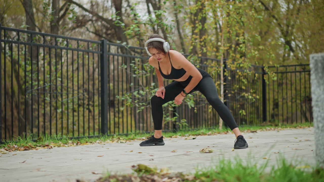 Young girl in black activewear stretching her legs outdoors, tapping her toe with hand while listening to music, blurred background showing iron fence and trees, enjoying fitness routine