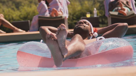 man on christmas holiday floating on inflatable ring in swimming pool wearing santa hat