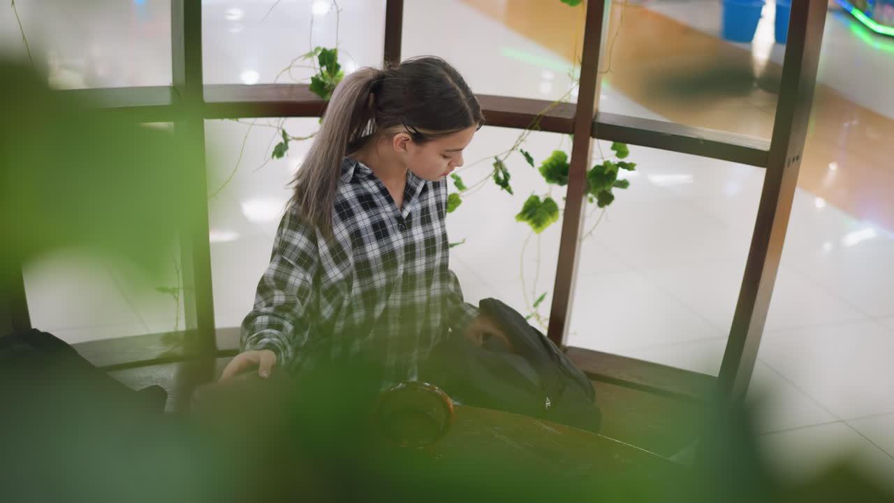 어린 소녀는 나무 의자에 앉아 가방에서 물건을 풀고, 흐릿한 녹색 꽃이 부분적으로 얼굴을 고, 자연 장식과 부드러운 조명으로 편안한 실내 환경은 평온한 분위기를 향상시니다.